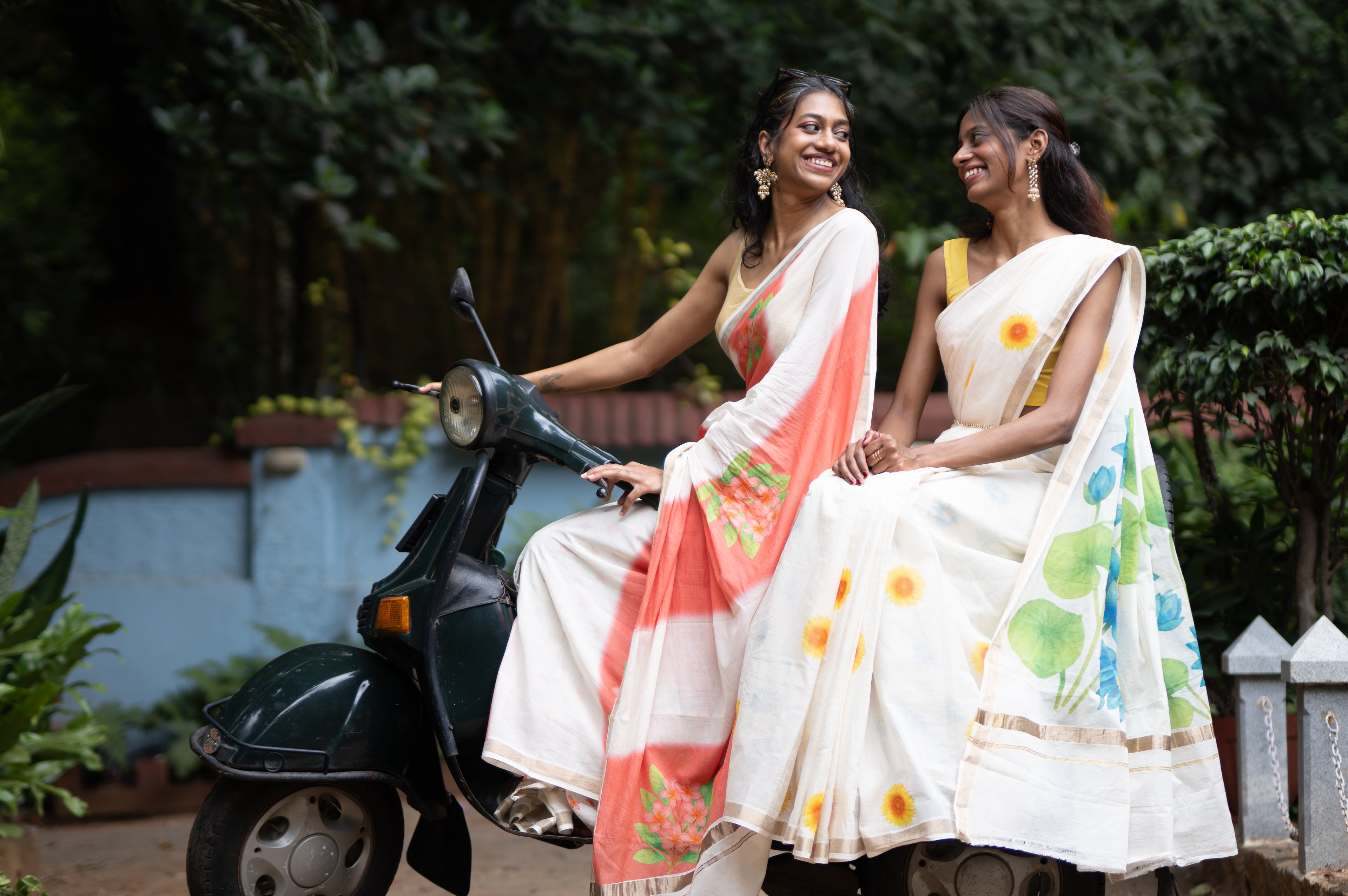 Two women in sarees sitting on a scooter with a greenery background